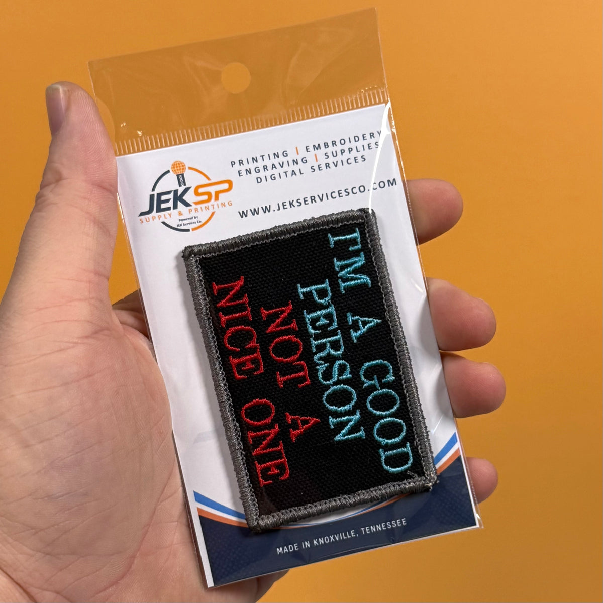 A hand holds a packaged embroidered patch against a solid orange background. The patch reads 'I'M A GOOD PERSON NOT A NICE ONE' in red, light blue, and white on a black background.