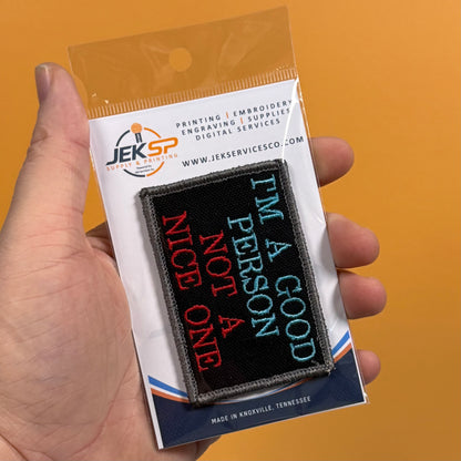 A hand holds a packaged embroidered patch against a solid orange background. The patch reads 'I'M A GOOD PERSON NOT A NICE ONE' in red, light blue, and white on a black background.