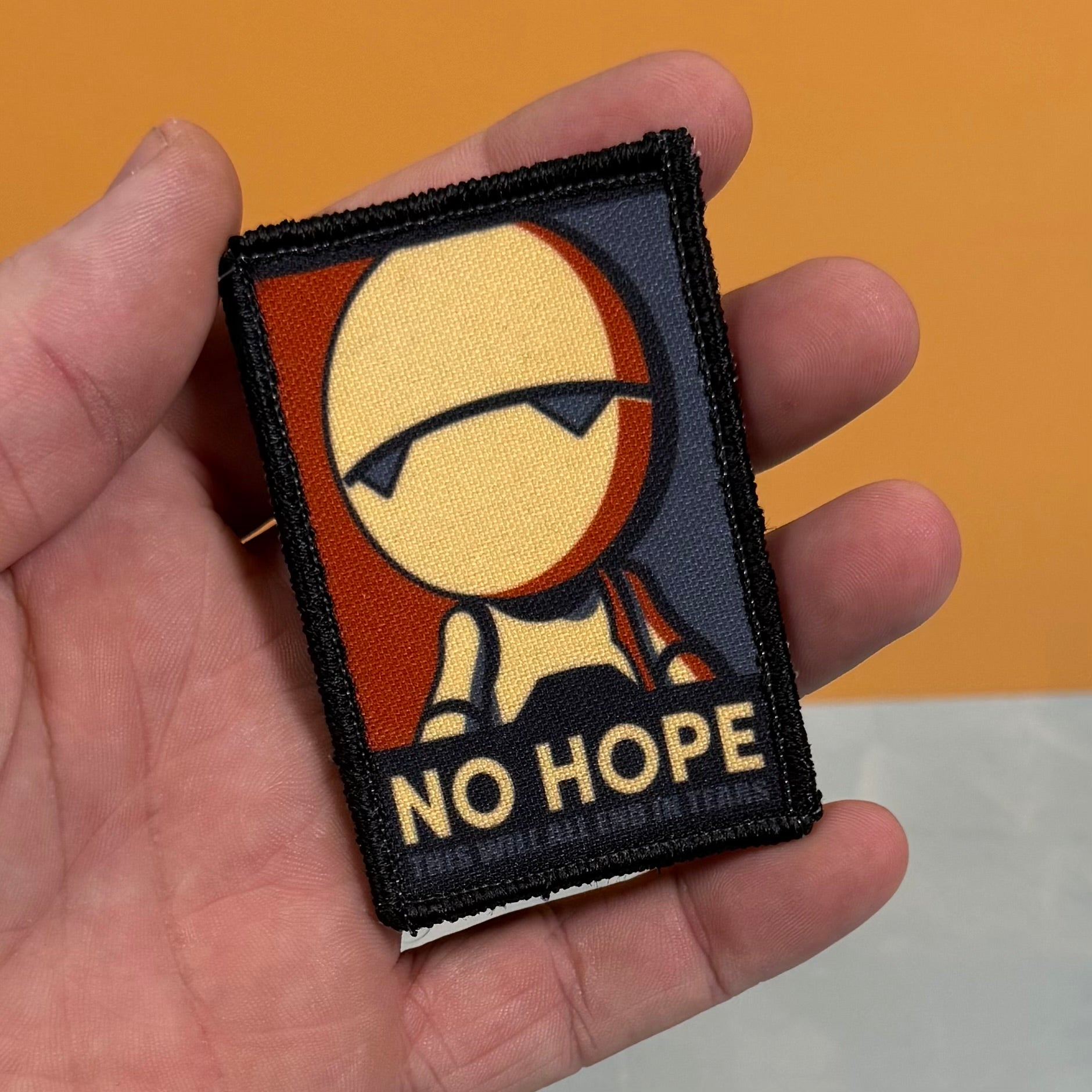 A hand holding a rectangular embroidered patch featuring a stylized face with sunglasses and the text 'NO HOPE' on a black background.