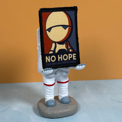 A small astronaut figurine holding a rectangular embroidered patch with a stylized face and the words “NO HOPE.” The figurine stands on a gray base against an orange wall and pale surface.