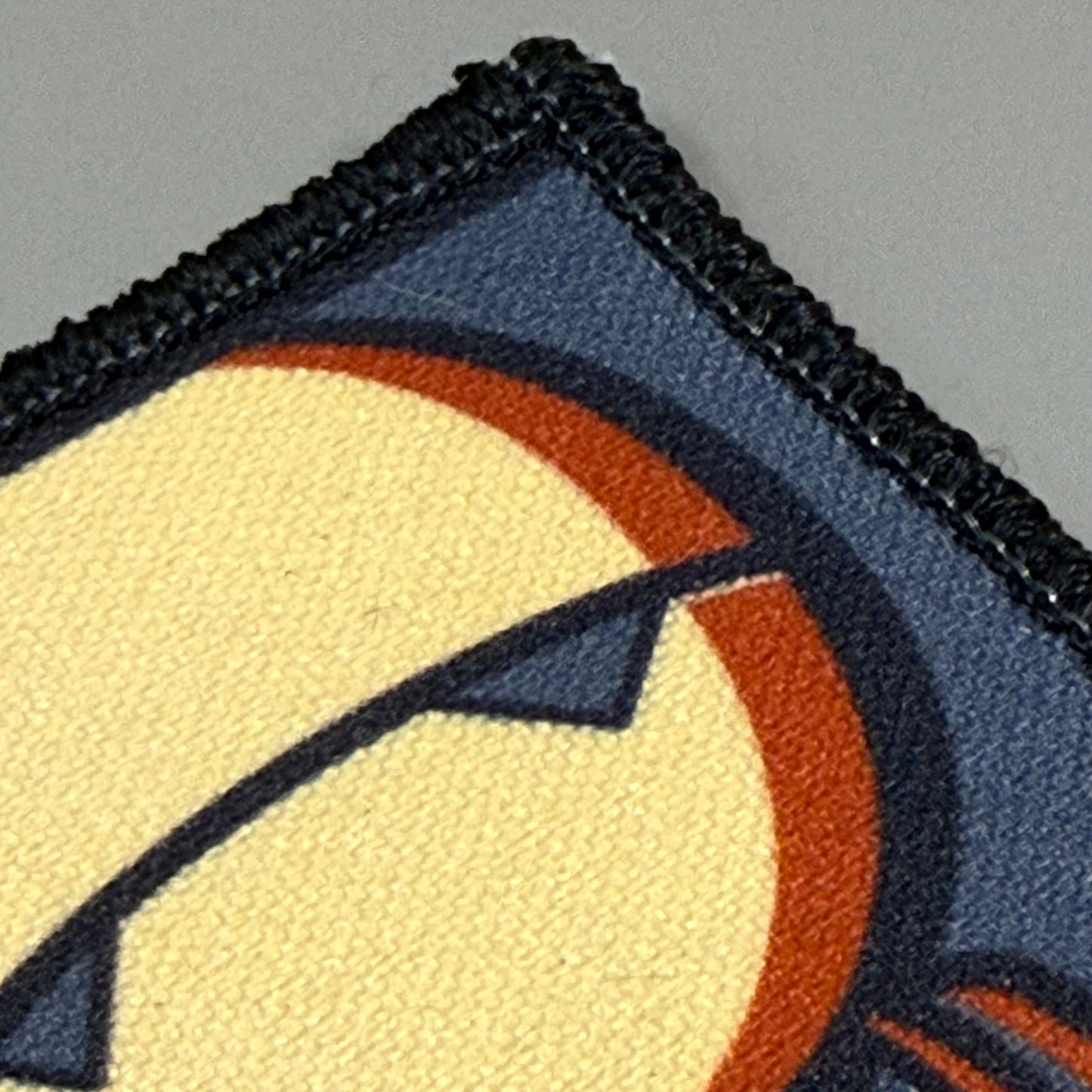 Close-up of a fabric patch showing part of a stylized face design in cream, dark blue, orange, and black, with a visible stitched border on a gray background.