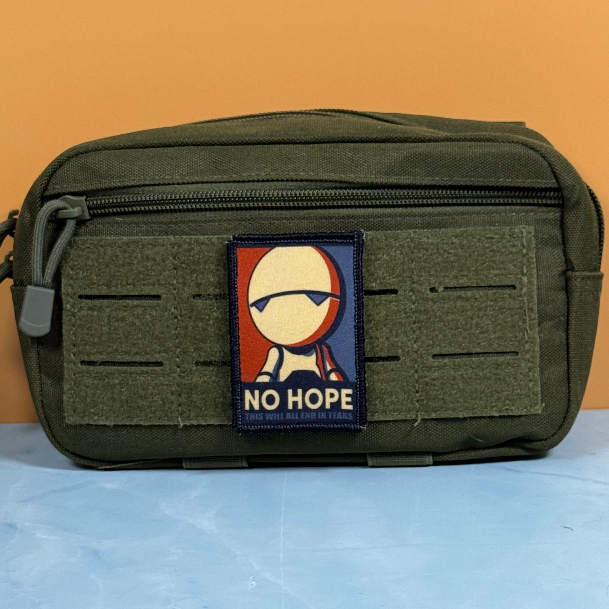 An olive green tactical pouch with a front zipper and hook-and-loop panel, featuring a rectangular 'NO HOPE' patch with a stylized figure on the front, set against a two-tone orange and blue background.