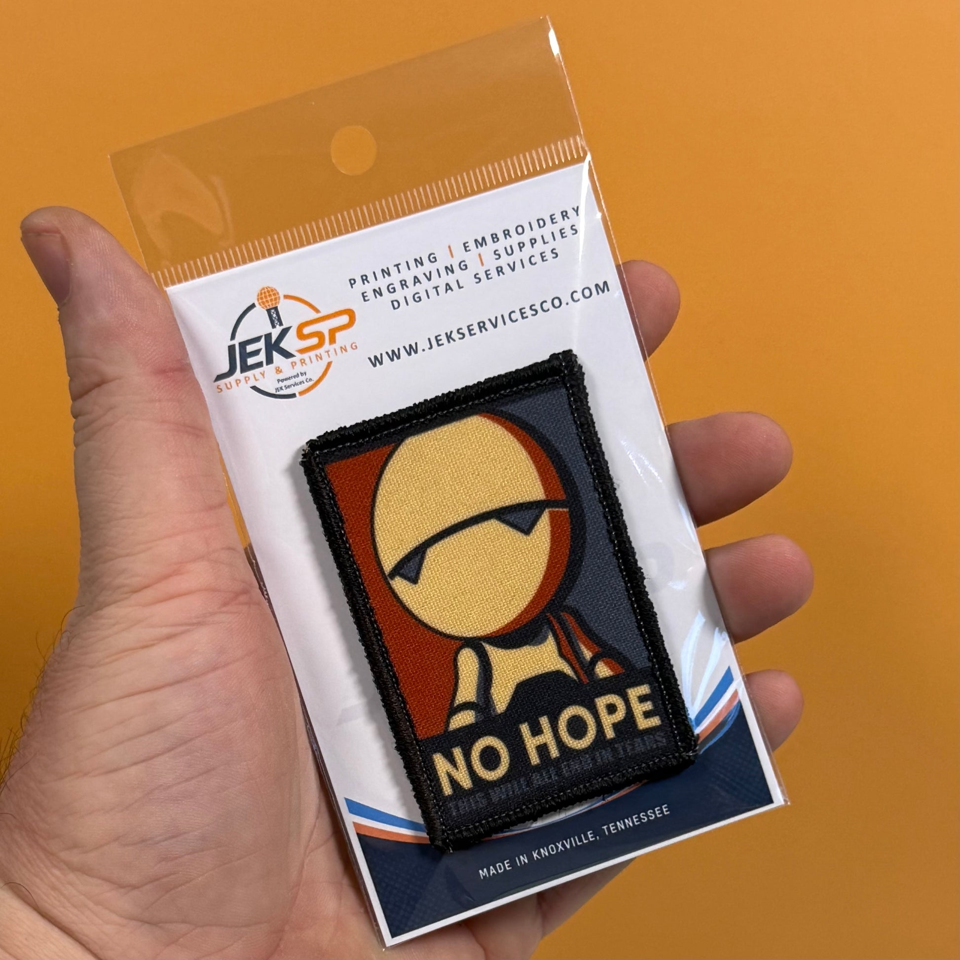 A hand holds a packaged embroidered patch against a solid orange background. The patch features a stylized figure with the text 'NO HOPE' and is mounted on a white backing card with the JEKSP brand logo.