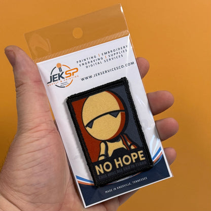 A hand holds a packaged embroidered patch against a solid orange background. The patch features a stylized figure with the text 'NO HOPE' and is mounted on a white backing card with the JEKSP brand logo.