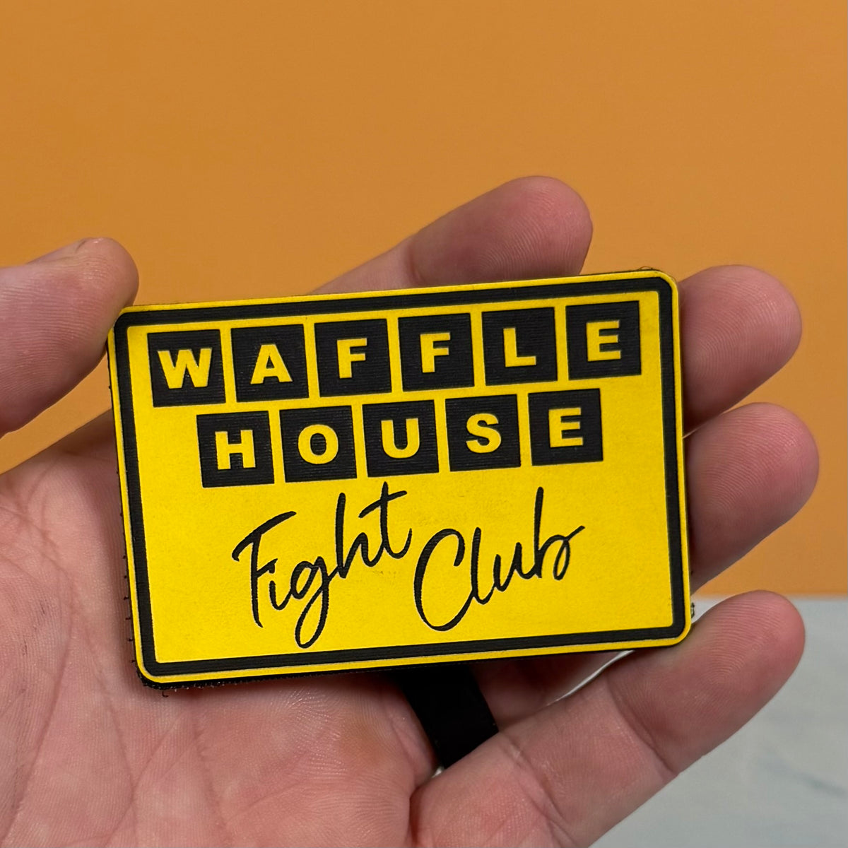 A hand holding a yellow rectangular patch with black text reading 'WAFFLE HOUSE Fight Club' against an orange background.