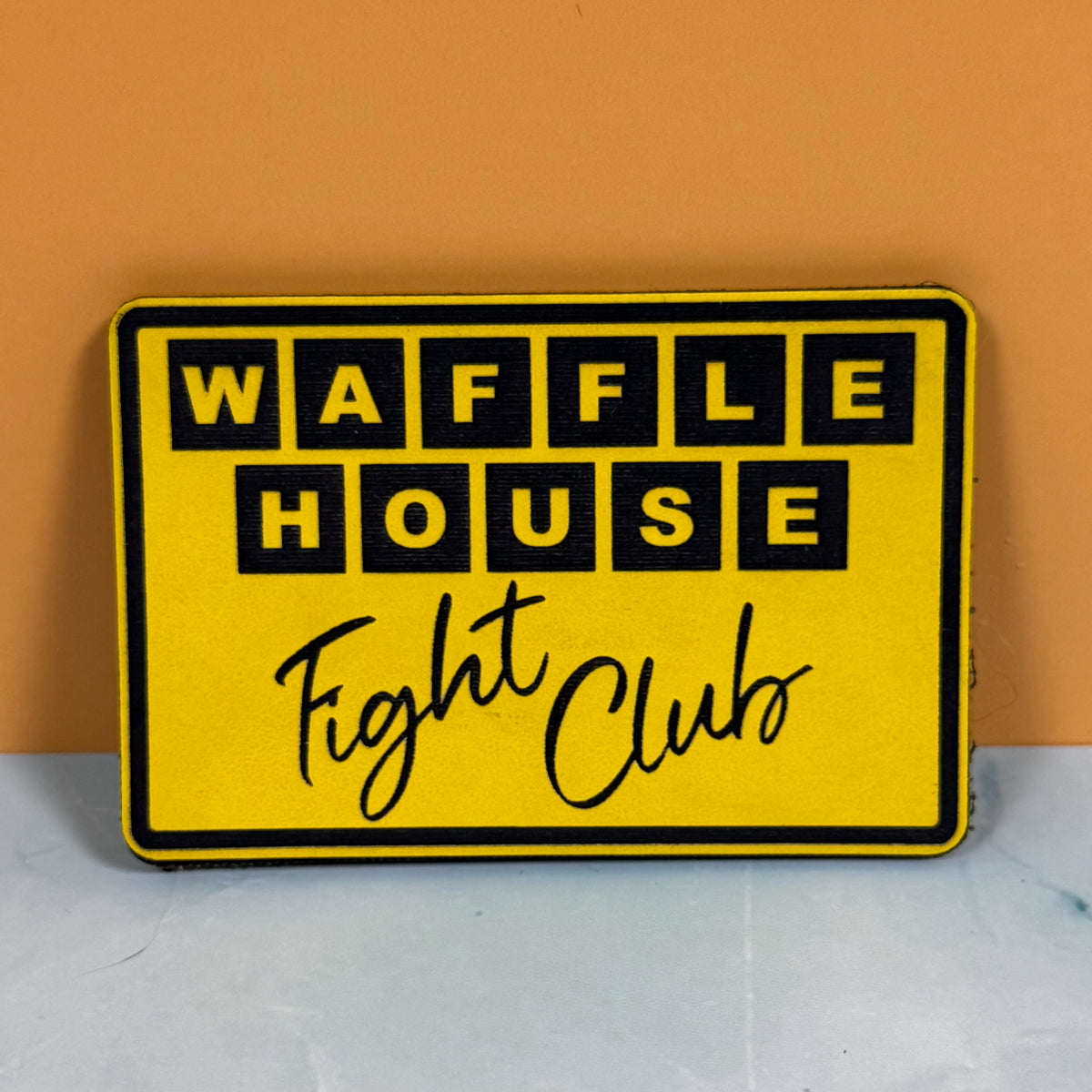 A yellow rectangular sign with a black border reading 'WAFFLE HOUSE Fight Club' in black lettering, placed against an orange wall and light gray surface.