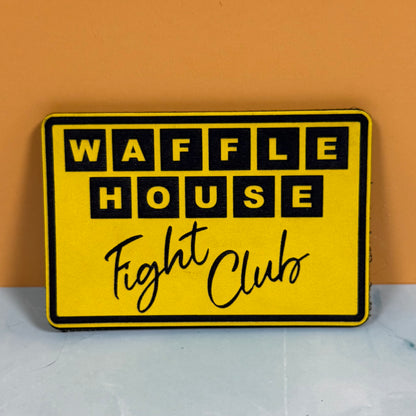A yellow rectangular sign with a black border reading 'WAFFLE HOUSE Fight Club' in black lettering, placed against an orange wall and light gray surface.