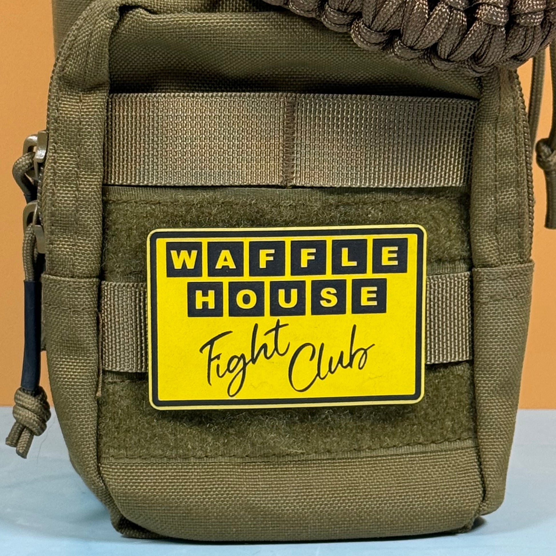 An olive green tactical pouch with a yellow and black 'WAFFLE HOUSE Fight Club' patch, shown against a solid orange background.