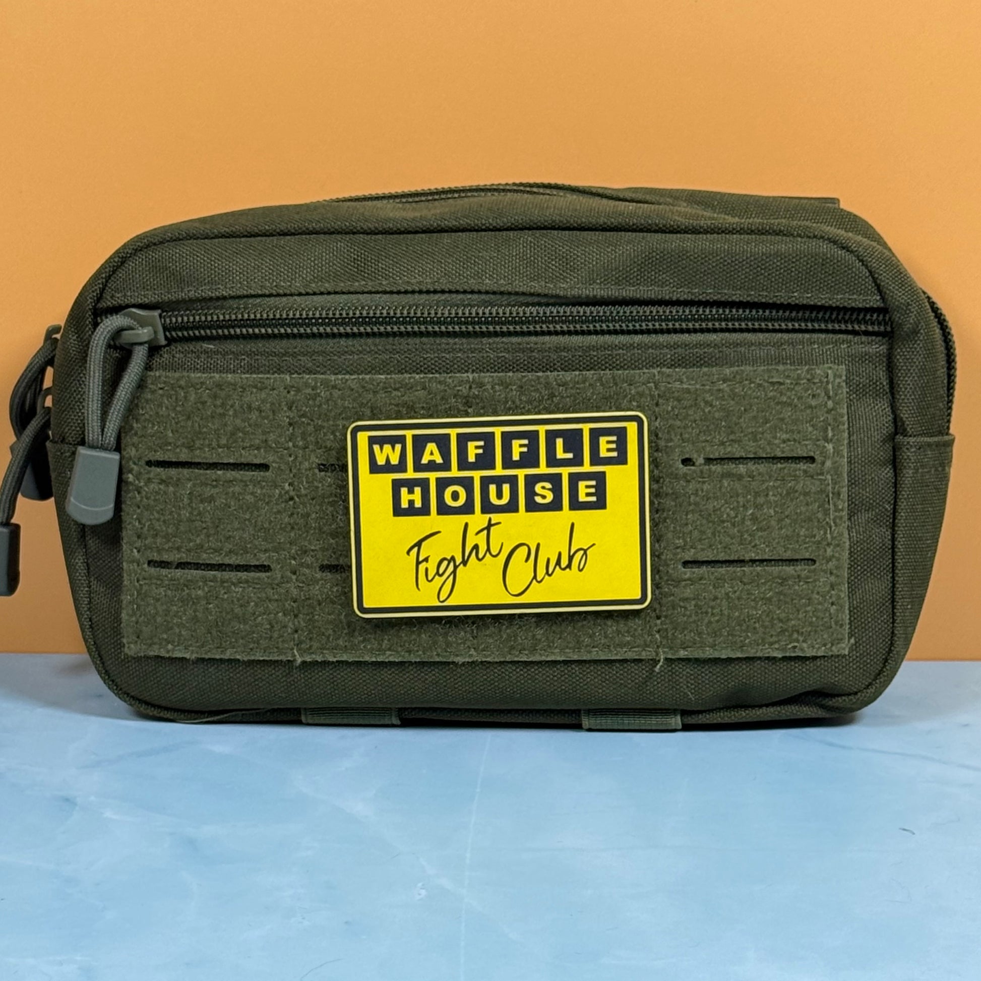 An olive green tactical pouch with a yellow 'WAFFLE HOUSE Fight Clubs' patch on the front, set against a solid orange background and light blue surface.