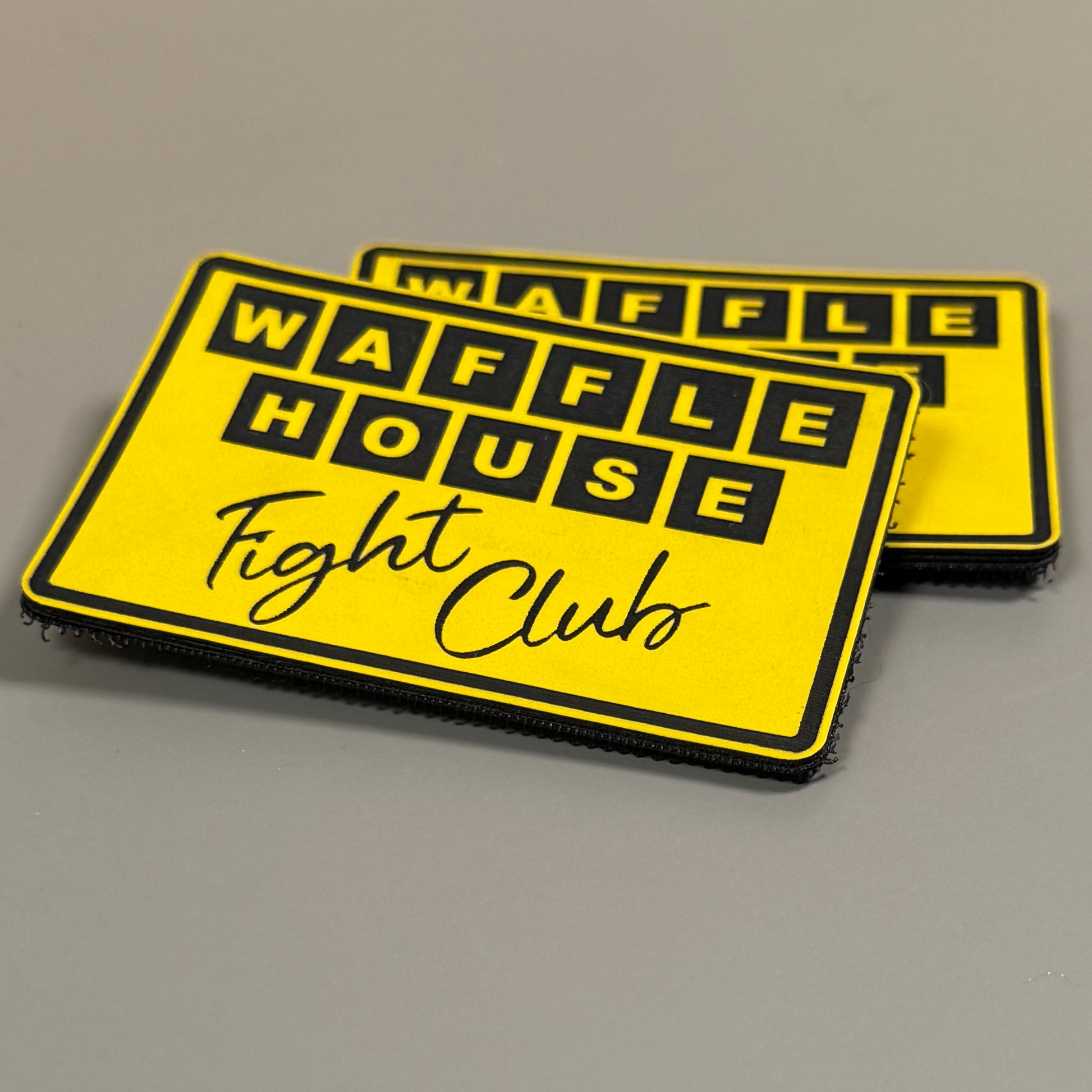 Two yellow rectangular patches with black borders and the text 'WAFFLE HOUSE Fight Club' in black, photographed on a plain gray surface.