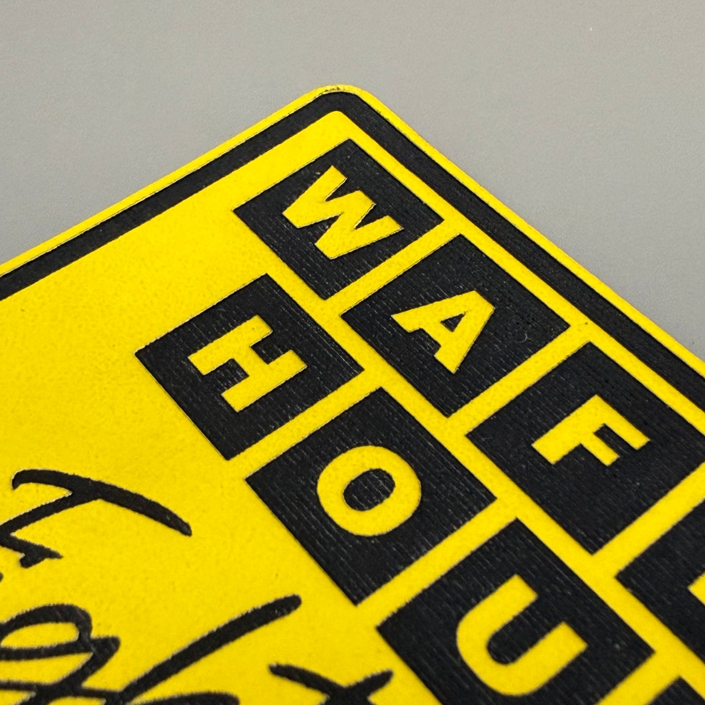 A close-up of a yellow sign or sticker with bold black text reading 'WAF HOU' inside black square blocks, photographed against a neutral gray background.