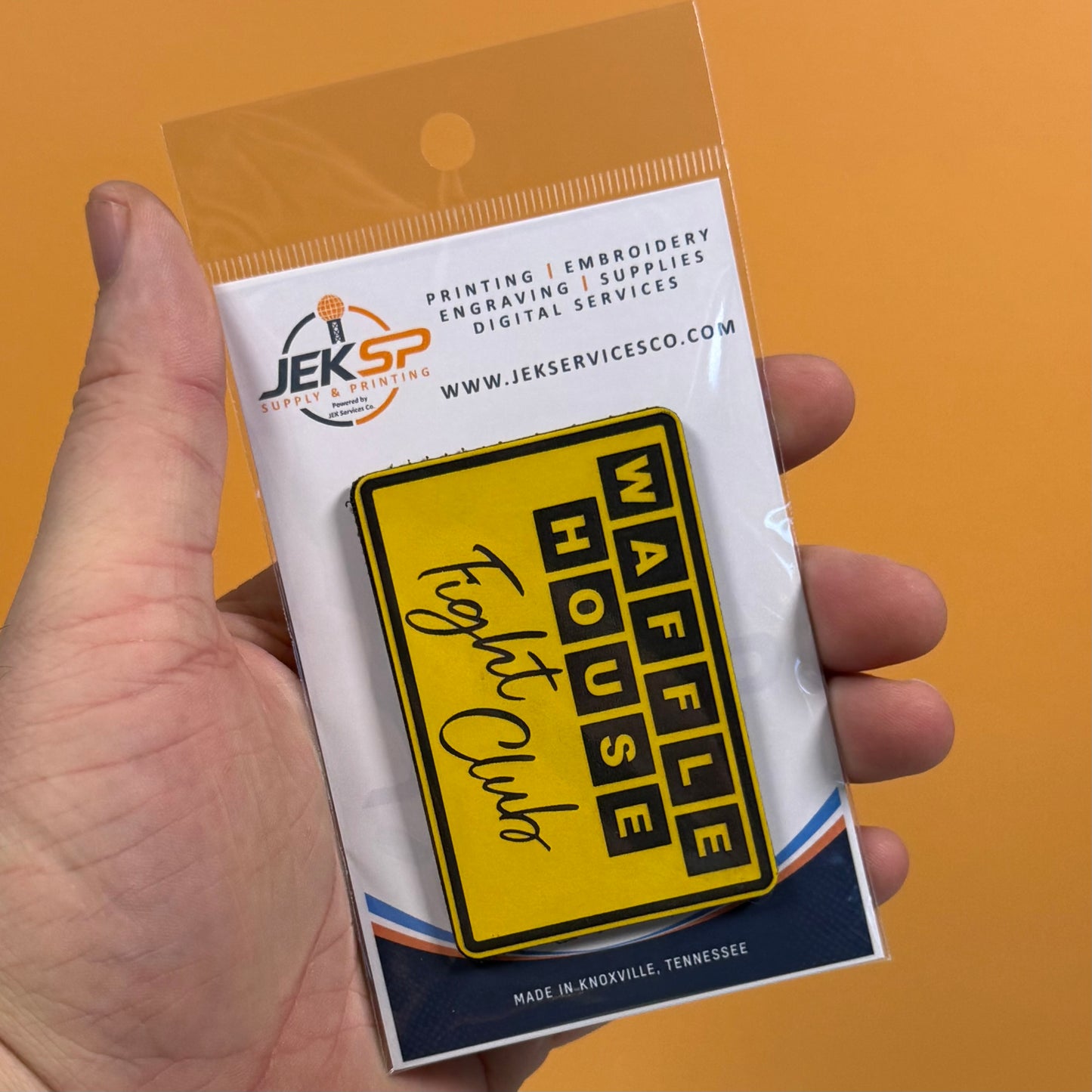 A hand holding a packaged yellow rectangular patch that reads 'WAFFLE HOUSE' and 'Fogey Club' on an orange background. The patch is attached to a white backing card with the Jeksp brand logo and text.