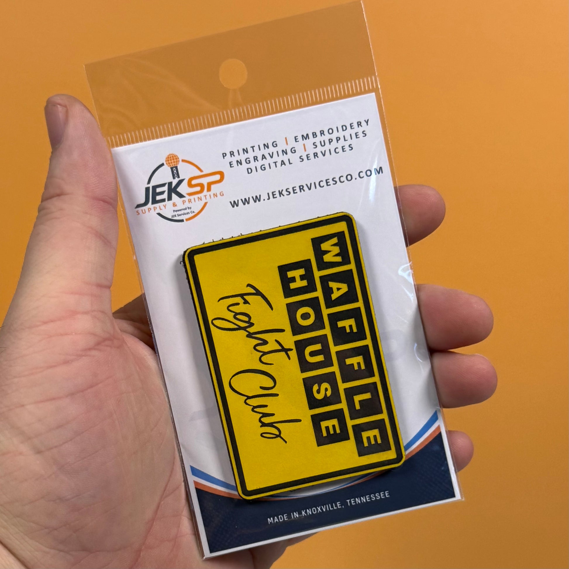 A hand holding a packaged yellow rectangular patch that reads 'WAFFLE HOUSE' and 'Fogey Club' on an orange background. The patch is attached to a white backing card with the Jeksp brand logo and text.