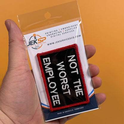 A hand holds a packaged embroidered patch that reads 'NOT THE WORST EMPLOYEE' in white letters on a black background with a red border, against an orange background. The package shows the brand JEKSP Design Systems.