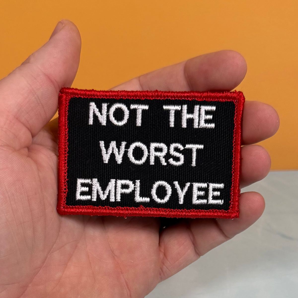 A hand holding a rectangular embroidered patch with the text 'NOT THE WORST EMPLOYEE' in white on a black background, bordered in red. The hand is positioned against a solid orange background.