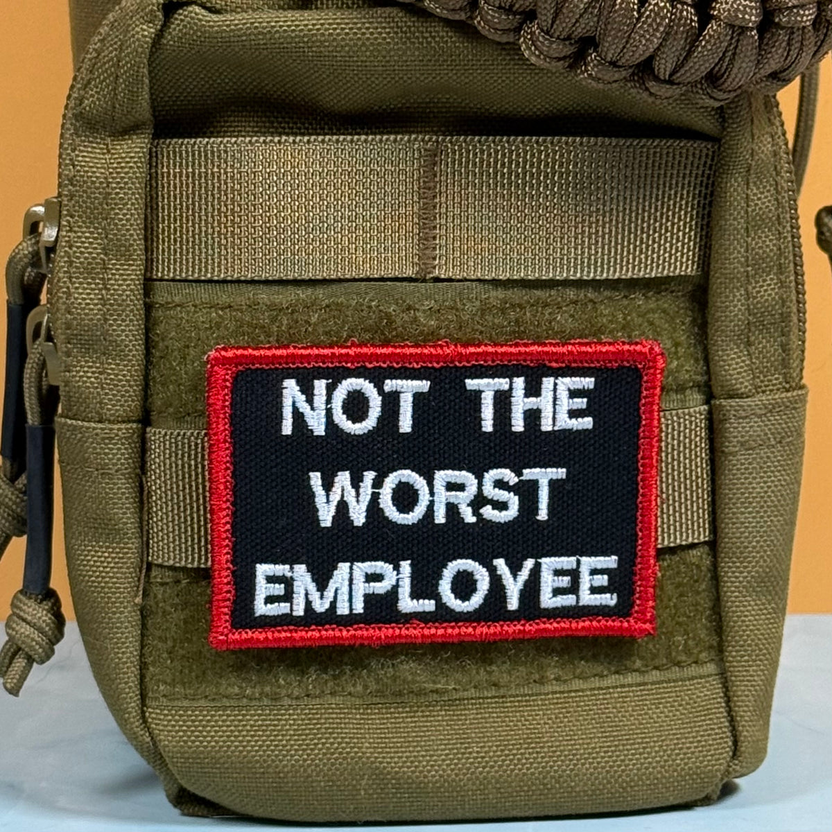 An olive green tactical pouch featuring a rectangular embroidered patch with the text 'NOT THE WORST EMPLOYEE' in white on a black background, bordered in red.
