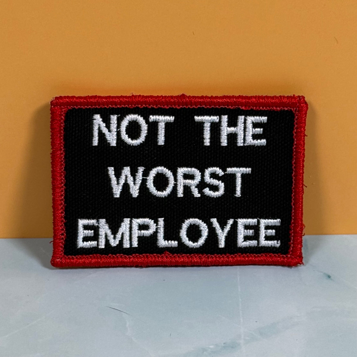 A rectangular embroidered patch with a black background, red border, and white text reading 'NOT THE WORST EMPLOYEE' against a two-tone orange and gray background.