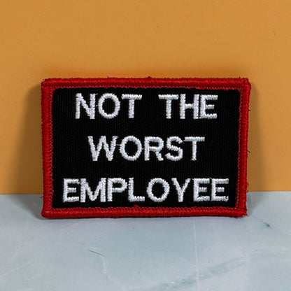 A rectangular embroidered patch with a black background, red border, and white text reading 'NOT THE WORST EMPLOYEE' against a two-tone orange and gray background.