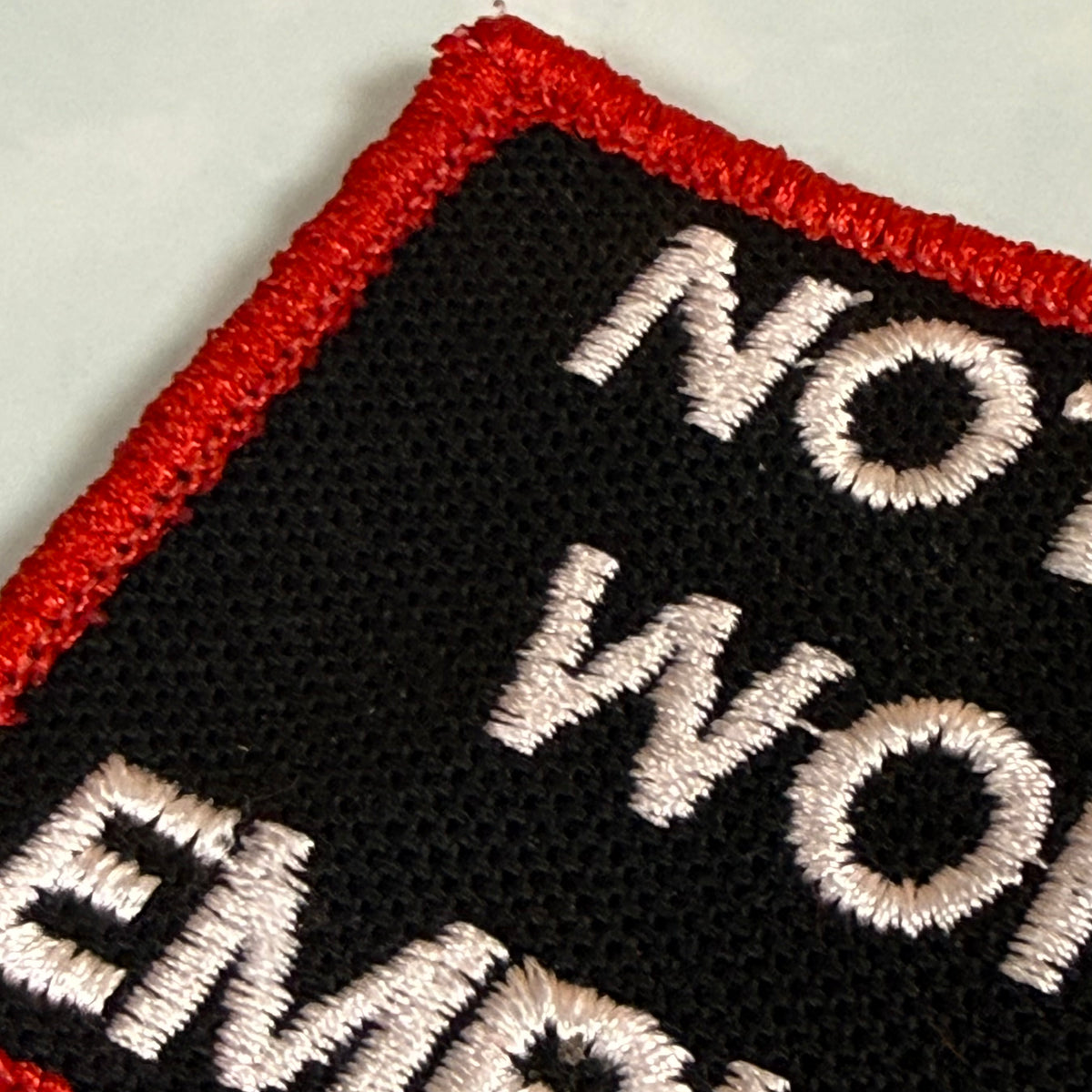 Close-up of an embroidered patch with a black background, red border, and white text reading 'NOT WORK EMPLOYMENT' on a light gray surface.