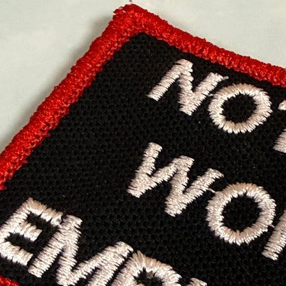 Close-up of an embroidered patch with a black background, red border, and white text reading 'NOT WORK EMPLOYMENT' on a light gray surface.