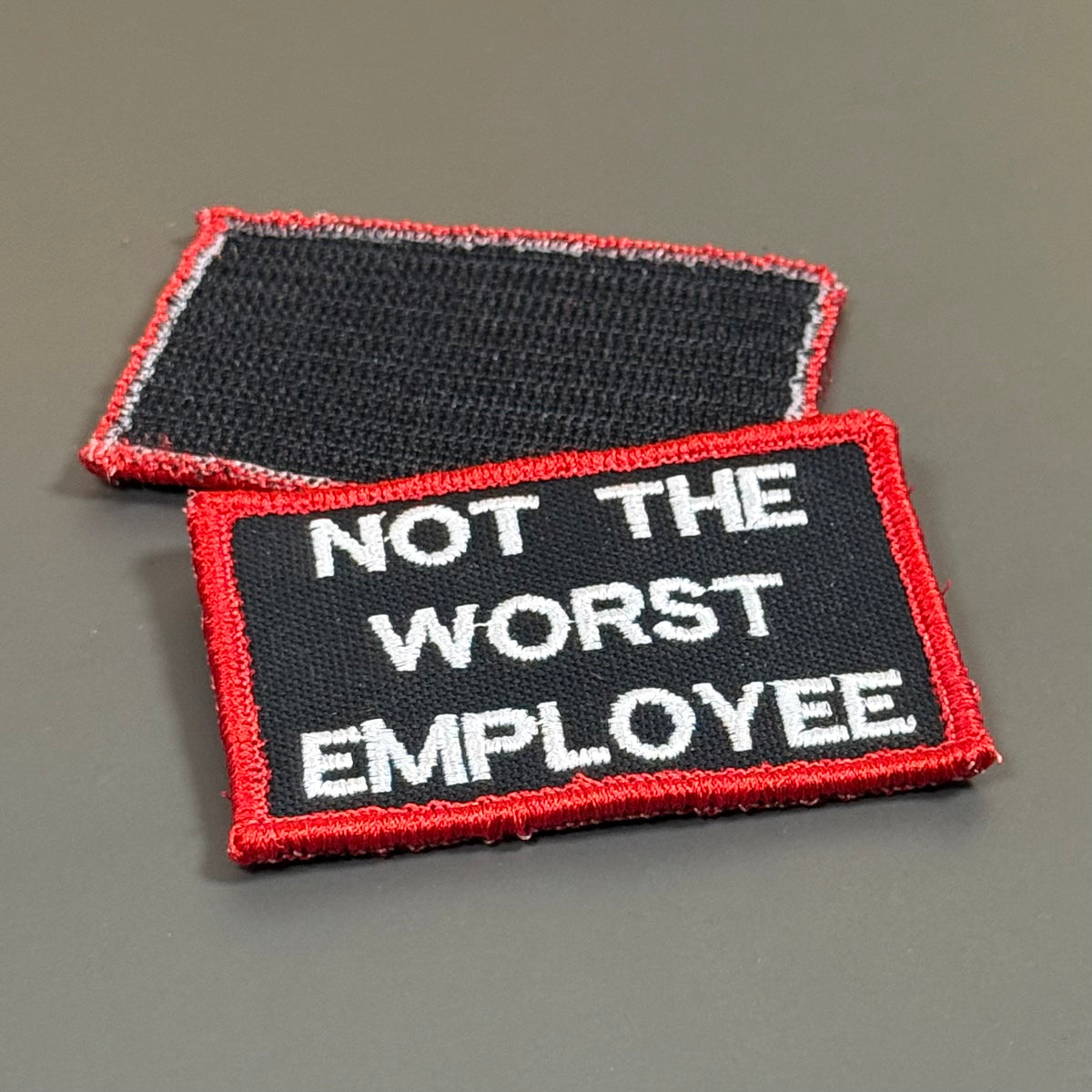 Two embroidered rectangular patches on a plain gray background. The front patch is black with white text reading 'NOT THE WORST EMPLOYEE' and a red border, while the back patch shows the dark adhesive side with a red and white edge.