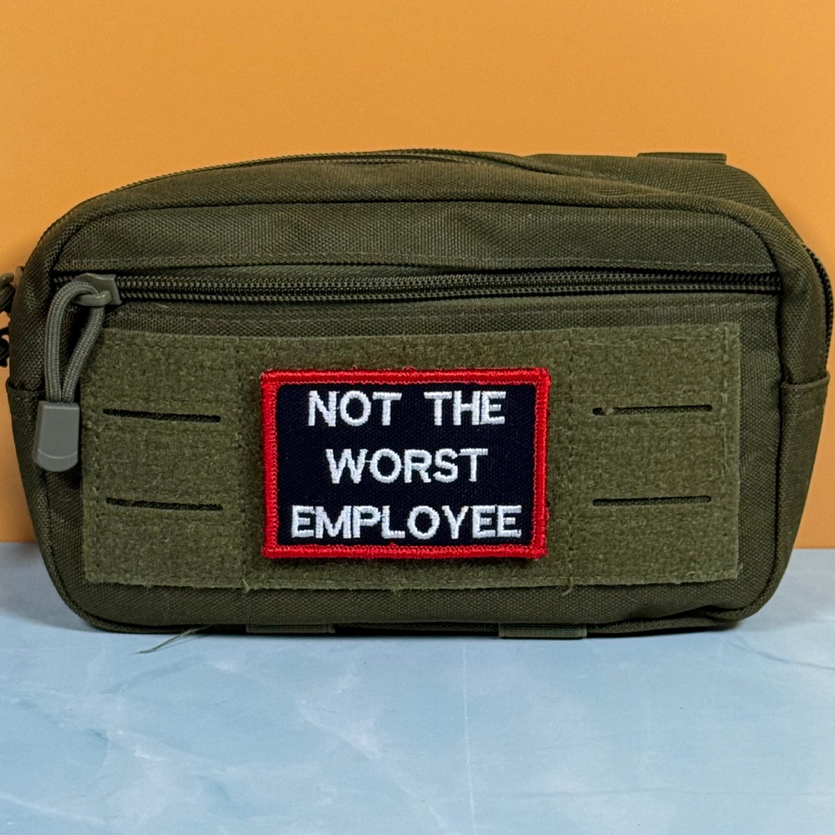 An olive green tactical pouch with a front zipper and a rectangular embroidered patch that reads 'NOT THE WORST EMPLOYEE' in white text on a black background with a red border, set against an orange and light blue background.