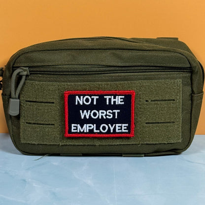 An olive green tactical pouch with a front zipper and a rectangular embroidered patch that reads 'NOT THE WORST EMPLOYEE' in white text on a black background with a red border, set against an orange and light blue background.