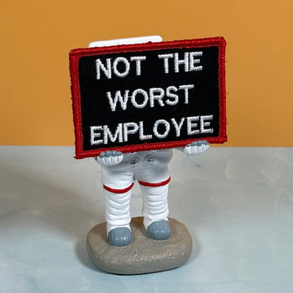 A novelty figurine of an astronaut in a white spacesuit holding a black sign with a red border that reads 'NOT THE WORST EMPLOYEE' in white letters, standing on a gray rock base against a two-tone orange and gray background.