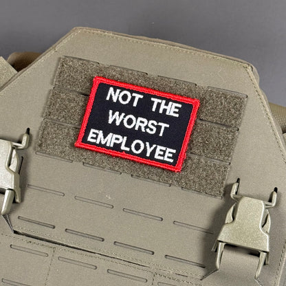 A rectangular embroidered patch with the text 'NOT THE WORST EMPLOYEE' in white on a black background with a red border, shown attached to a tan tactical plate carrier.