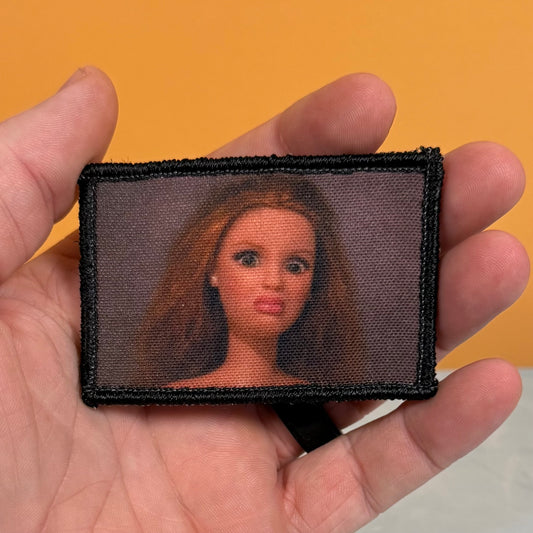 A rectangular embroidered patch with a black border and a printed portrait of a doll with long light brown hair, held in a person's hand against a solid orange background.