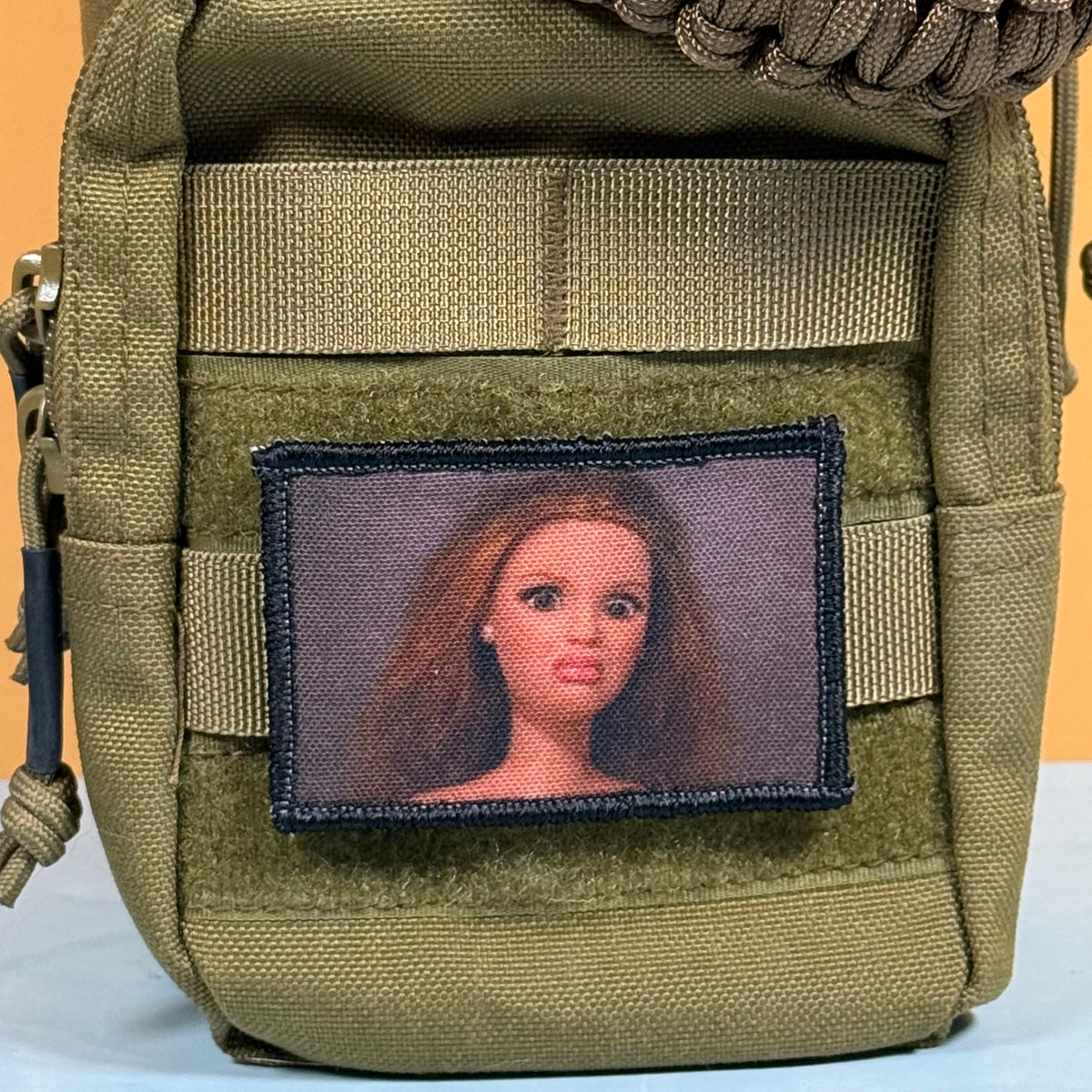 An olive green tactical pouch with MOLLE webbing and a hook-and-loop patch area, featuring a rectangular patch with a portrait of a doll with brown hair.