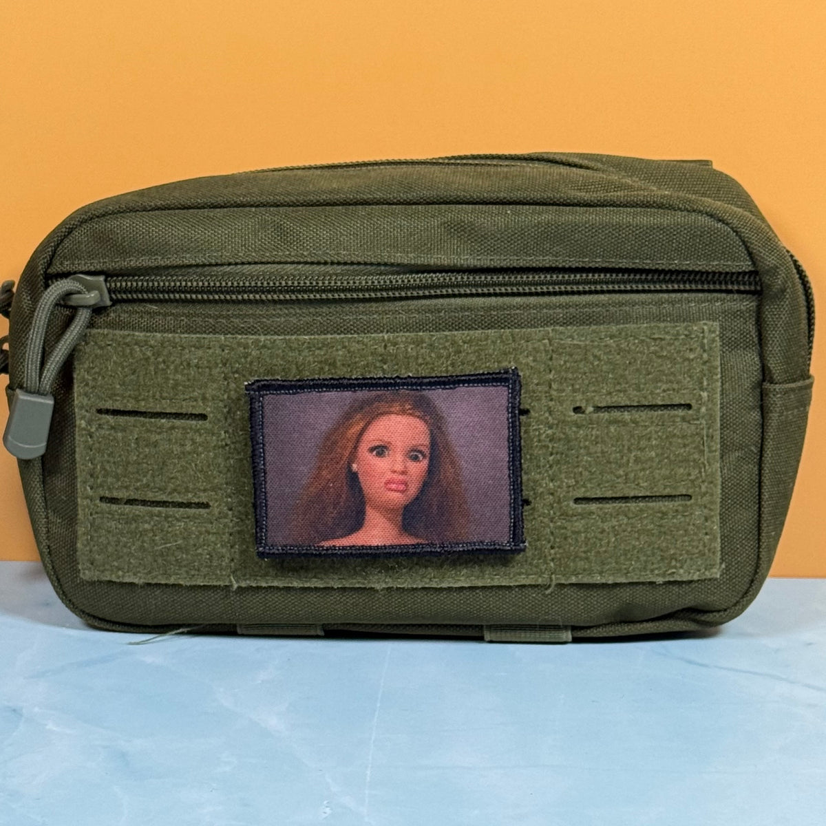 An olive green tactical pouch with a front zipper and a custom rectangular patch featuring a portrait of a woman with long light brown hair, set against a solid orange background.