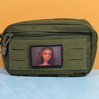 An olive green tactical pouch with a front zipper and a custom rectangular patch featuring a portrait of a woman with long light brown hair, set against a solid orange background.