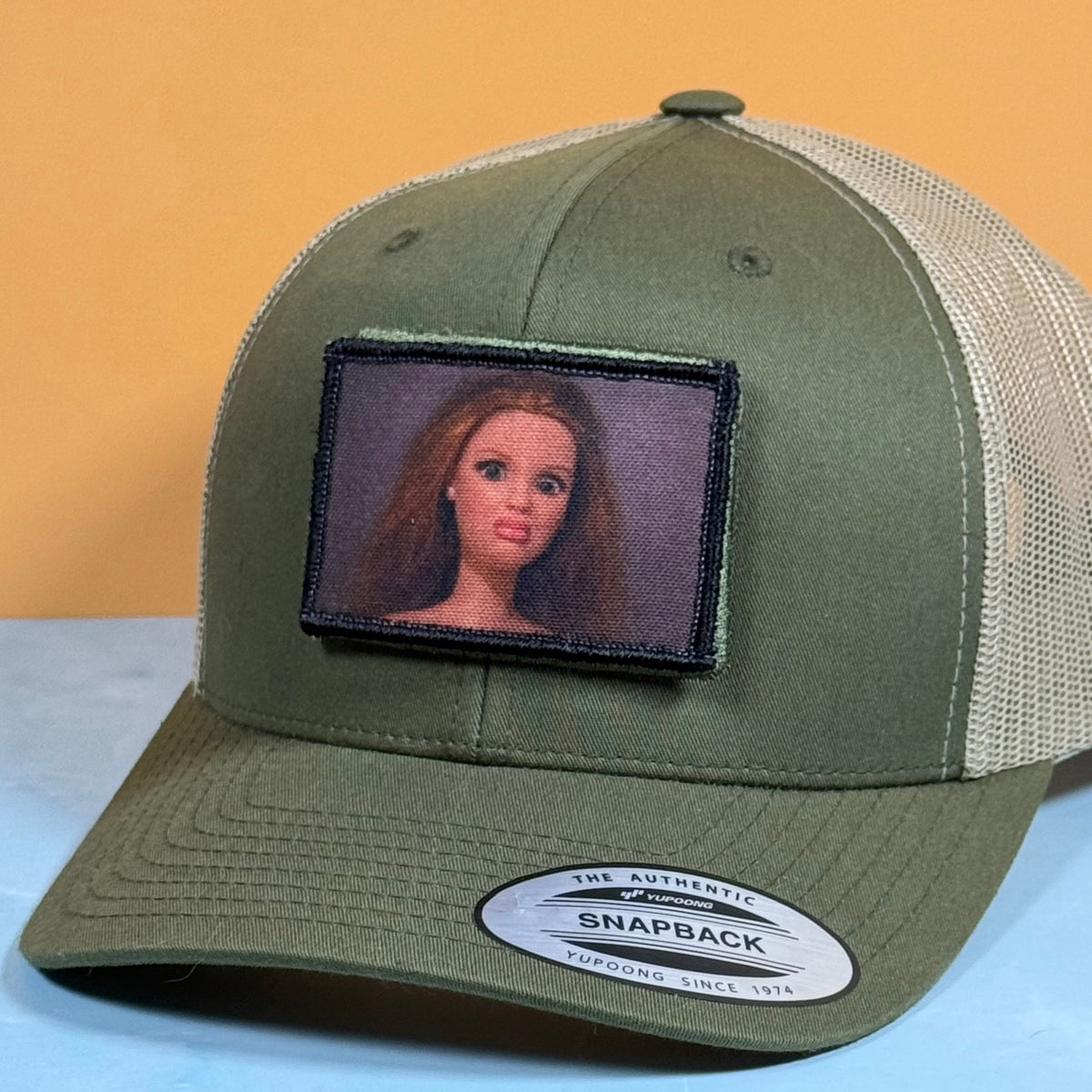 An olive green and beige mesh trucker hat with a custom embroidered patch of a doll's face on the front. The hat features a curved brim with a visible Snapback sticker and is photographed against a split yellow and light blue background.