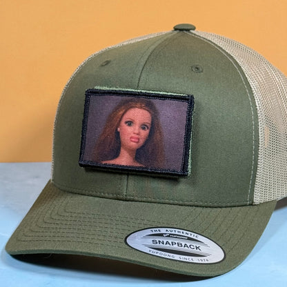 An olive green and beige mesh trucker hat with a custom embroidered patch of a doll's face on the front. The hat features a curved brim with a visible Snapback sticker and is photographed against a split yellow and light blue background.