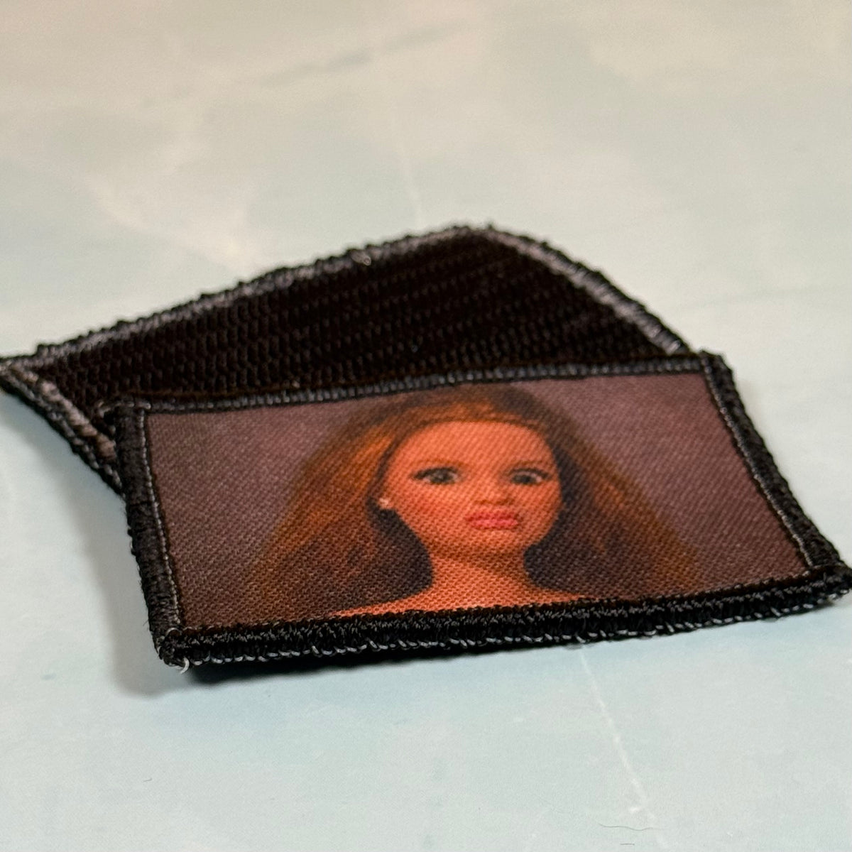 A rectangular embroidered patch with a portrait of a doll-like figure with long brown hair, shown on a light gray background with a second patch partially visible behind it.