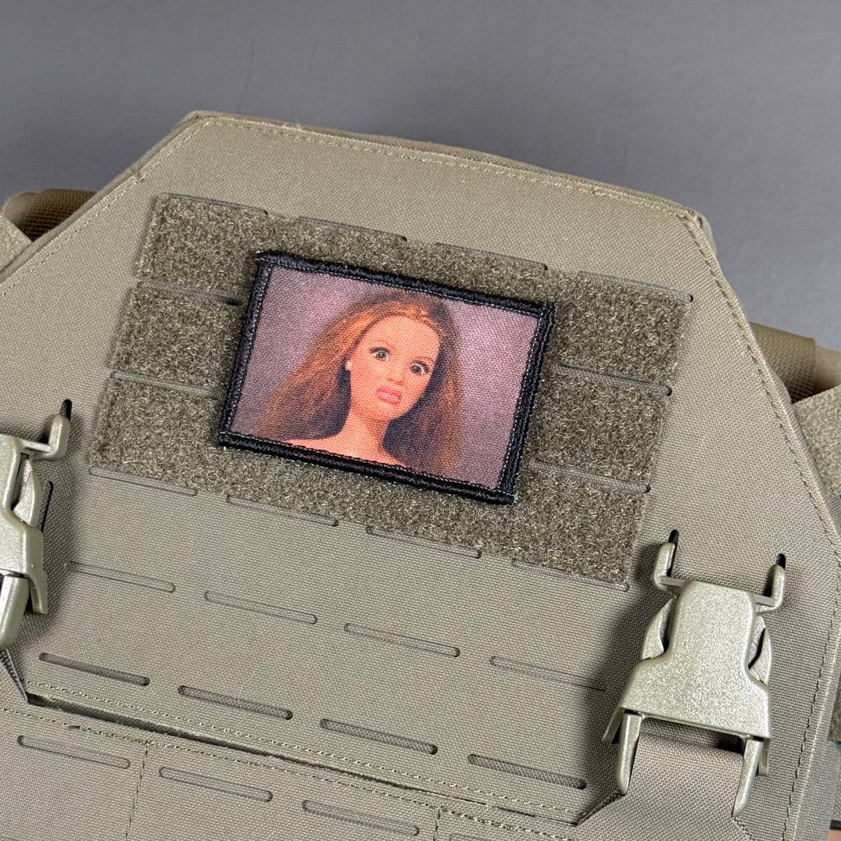A close-up of a tactical vest or plate carrier in tan/khaki fabric, featuring a rectangular embroidered patch with a portrait of a woman with long light brown hair on a gray background.