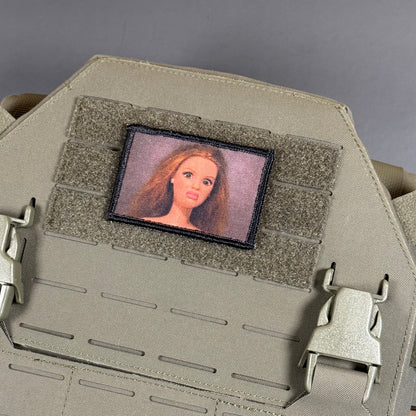 A close-up of a tactical vest or plate carrier in tan/khaki fabric, featuring a rectangular embroidered patch with a portrait of a woman with long light brown hair on a gray background.