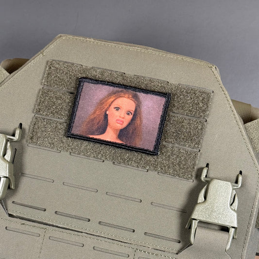 A close-up of a tactical vest or plate carrier in tan/khaki fabric, featuring a rectangular embroidered patch with a portrait of a woman with long light brown hair on a gray background.
