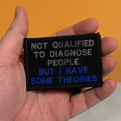 A hand holding a rectangular black embroidered patch with the text 'NOT QUALIFIED TO DIAGNOSE PEOPLE BUT I HAVE SOME THEORIES' in white and blue lettering, set against a solid orange background.