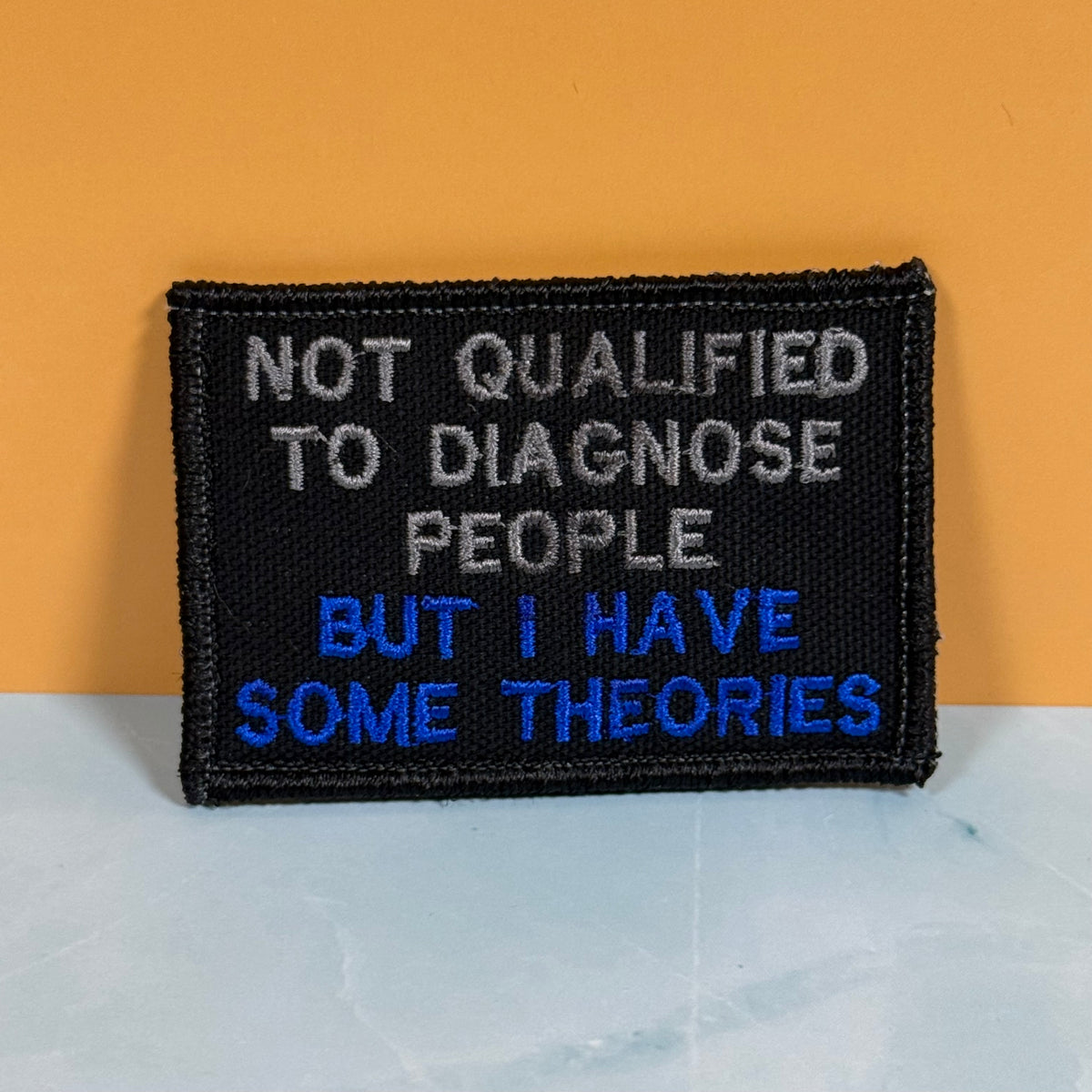 A rectangular black embroidered patch with the text 'NOT QUALIFIED TO DIAGNOSE PEOPLE BUT I HAVE SOME THEORIES' in white and blue lettering, displayed against a split orange and light gray background.
