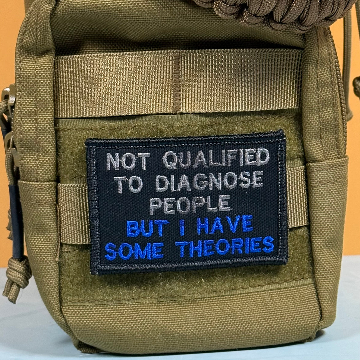 A close-up of a tactical pouch in olive drab/khaki nylon with a black embroidered patch that reads 'NOT QUALIFIED TO DIAGNOSE PEOPLE BUT I HAVE SOME THEORIES' in white and blue text.