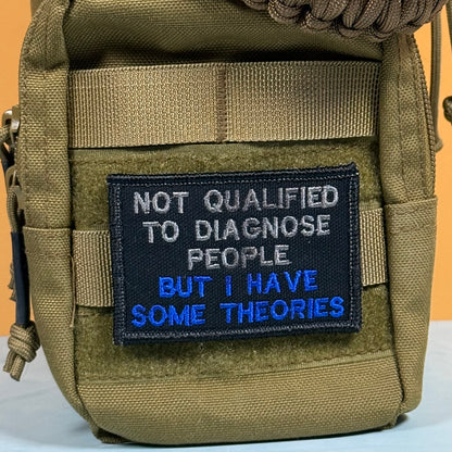 A close-up of a tactical pouch in olive drab/khaki nylon with a black embroidered patch that reads 'NOT QUALIFIED TO DIAGNOSE PEOPLE BUT I HAVE SOME THEORIES' in white and blue text.