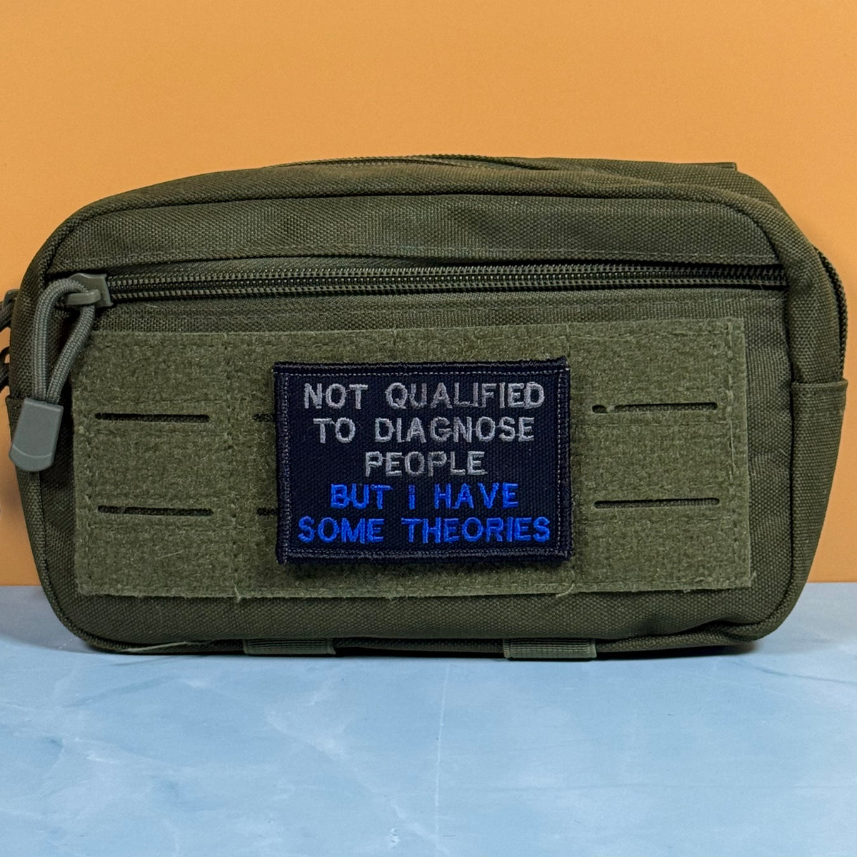 An olive green tactical pouch with a front zipper and a rectangular hook-and-loop patch. The patch is navy blue with white and blue embroidered text that reads, 'NOT QUALIFIED TO DIAGNOSE PEOPLE BUT I HAVE SOME THEORIES.