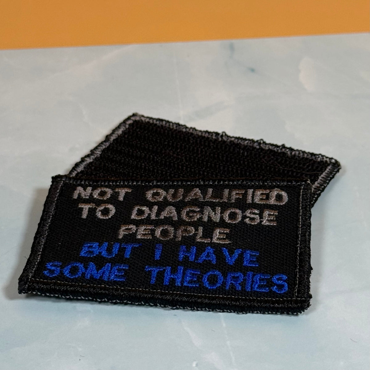 A rectangular embroidered patch with a black background and white border, featuring the text 'NOT QUALIFIED TO DIAGNOSE PEOPLE BUT I HAVE SOME THEORIES' in white and blue lettering, placed on a light marble-like surface.