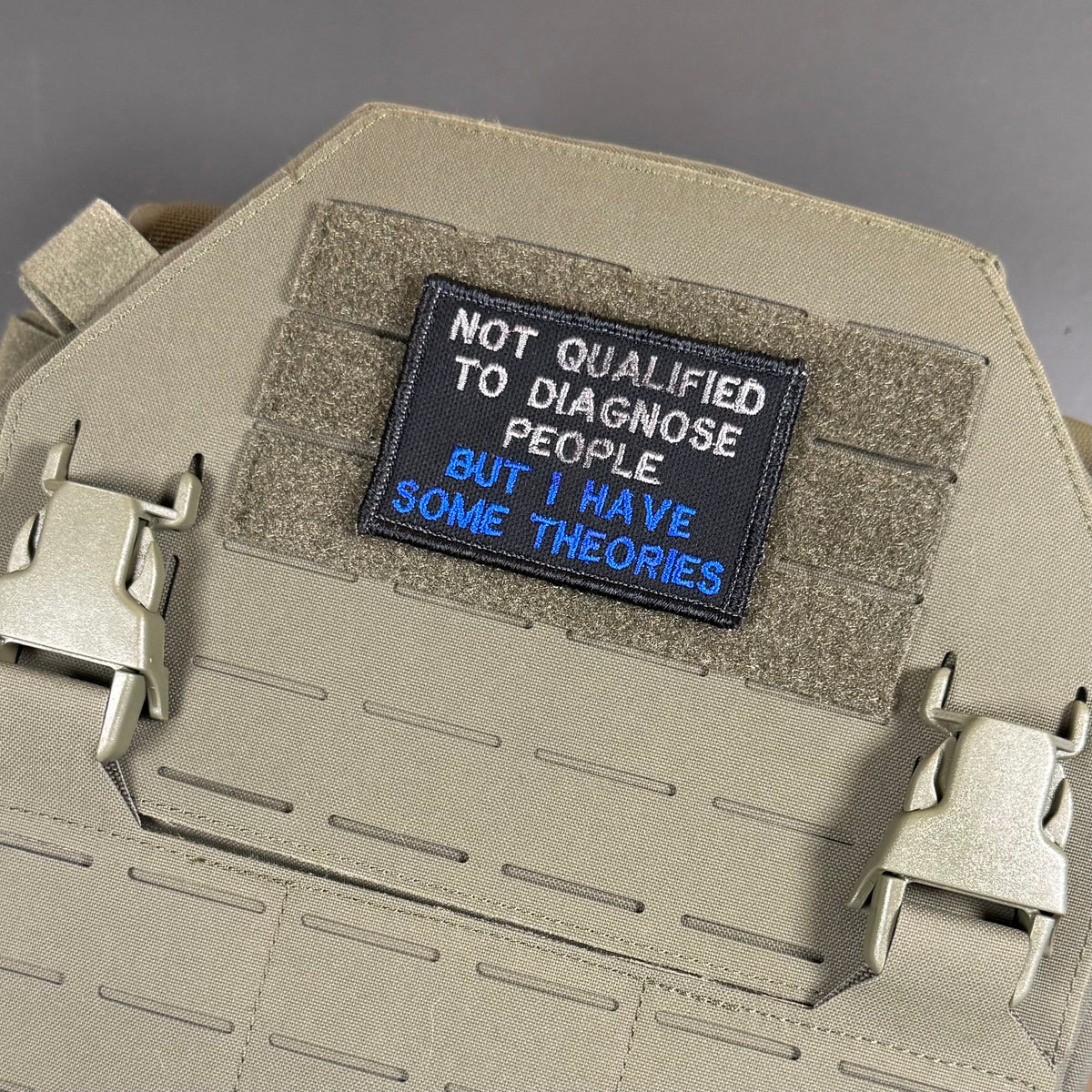 A tactical patch with the text 'NOT QUALIFIED TO DIAGNOSE PEOPLE. BUT I HAVE SOME THEORIES' in white and blue lettering on a black background, displayed on a tan MOLLE plate carrier vest.
