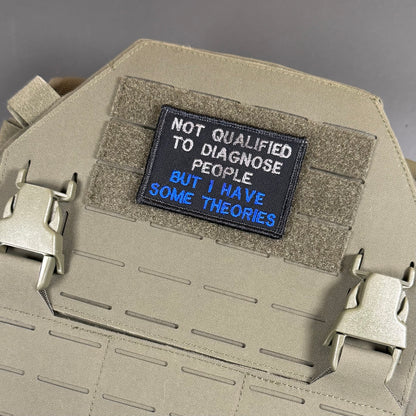 A tactical patch with the text 'NOT QUALIFIED TO DIAGNOSE PEOPLE. BUT I HAVE SOME THEORIES' in white and blue lettering on a black background, displayed on a tan MOLLE plate carrier vest.