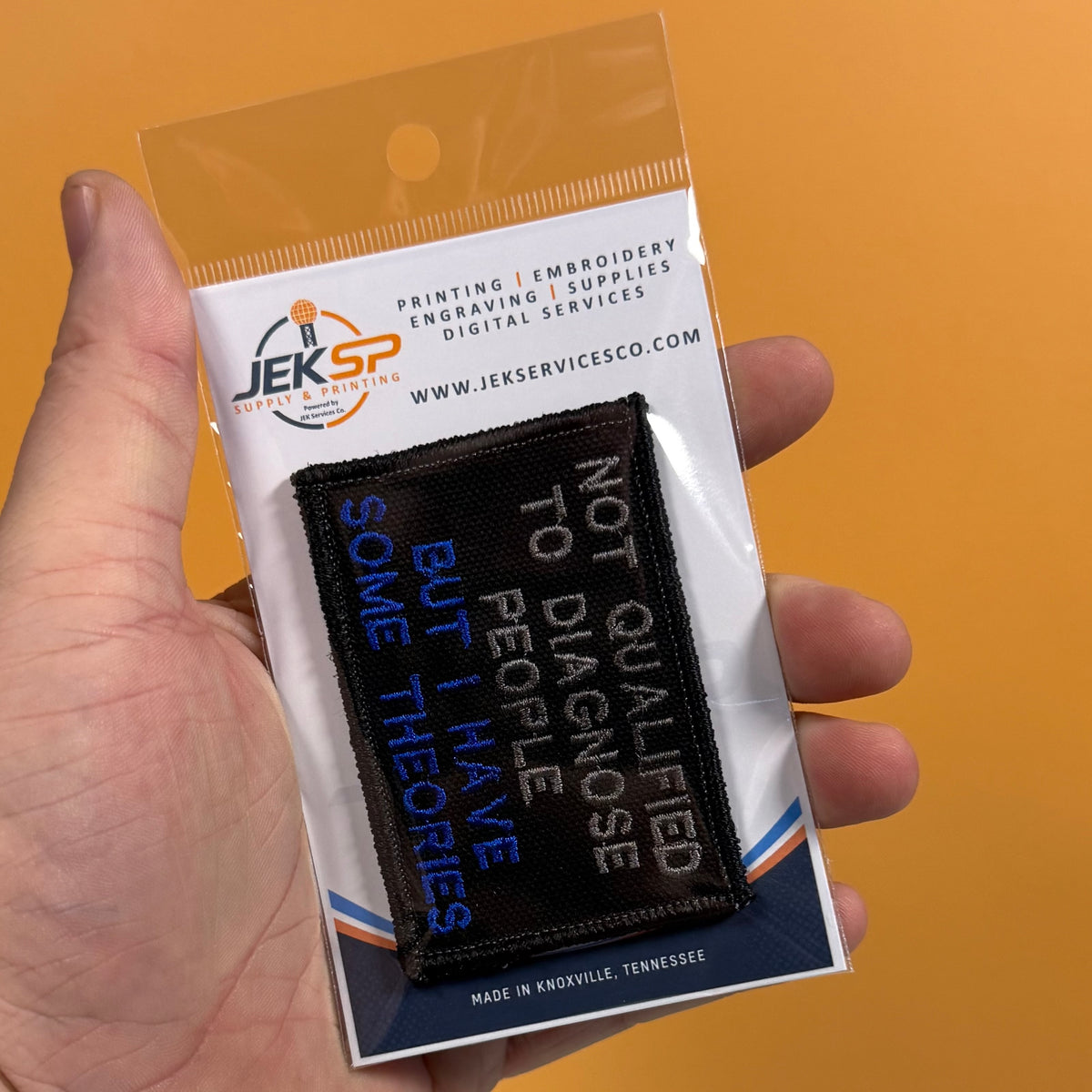 A hand holds a small embroidered patch in its retail packaging against a solid orange background. The patch is black with white and blue text reading 'NOT QUALIFIED TO DIAGNOSE SOME THEORIES BUT I HAVE'.