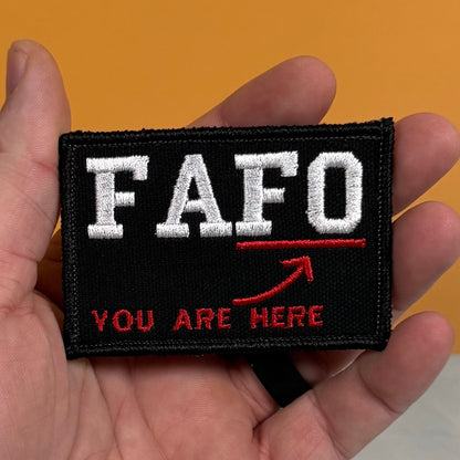 A close-up of a hand holding a rectangular black embroidered patch with the text 'FAFO' in white and 'YOU ARE HERE' in red, plus a red arrow, against an orange background.