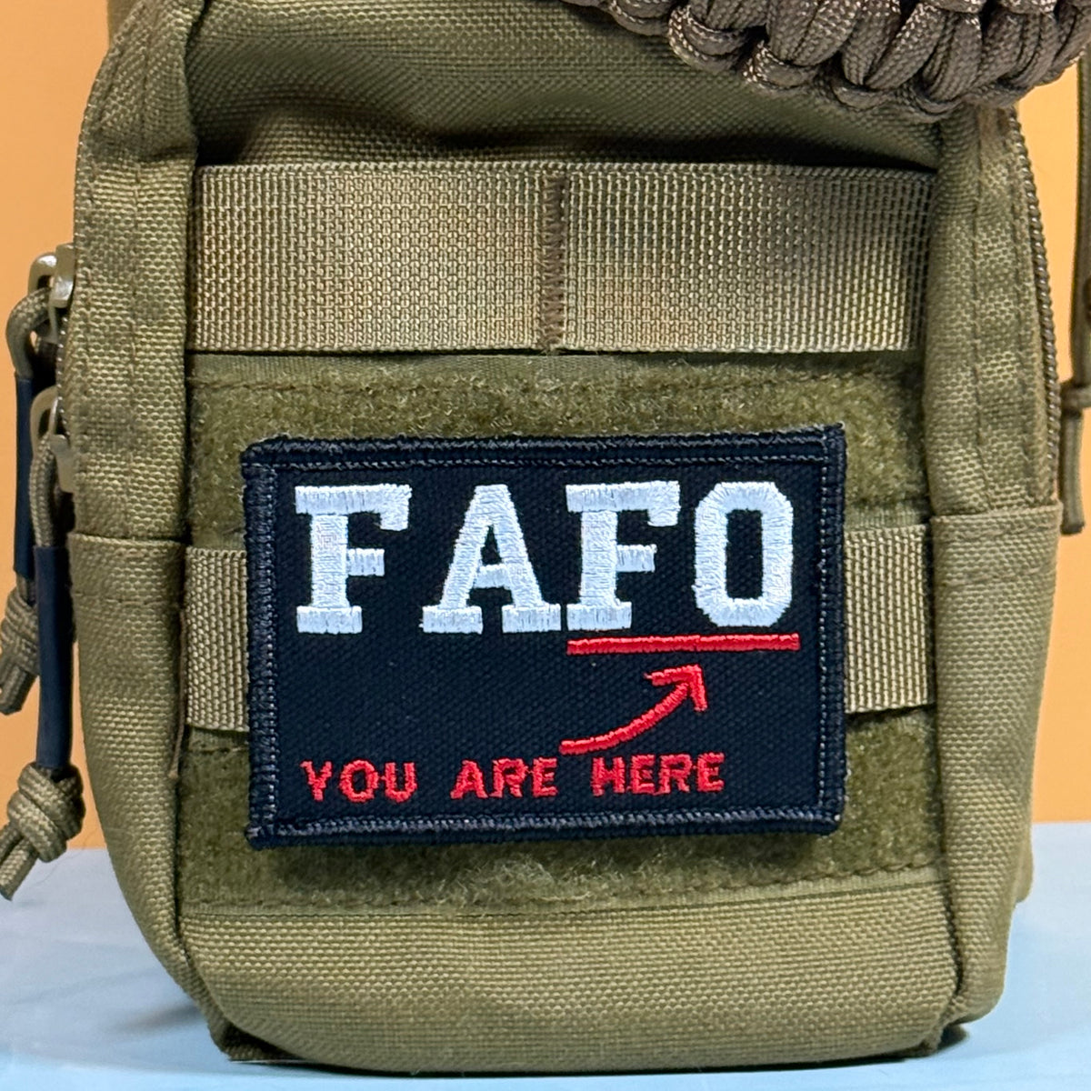 A close-up of a tactical utility pouch in olive drab green nylon, featuring a rectangular embroidered patch on the front. The patch is dark navy blue with the letters 'FAFO' in white, a red underline, a red arrow, and the words 'YOU ARE HERE' in red.